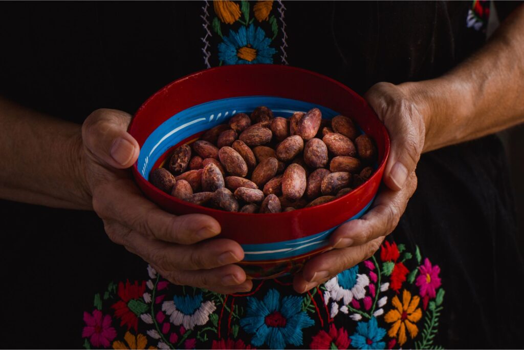 Woman with a traditional and colorful embroided dress holding a red and blue ceramic bowl filled with cacao beans.