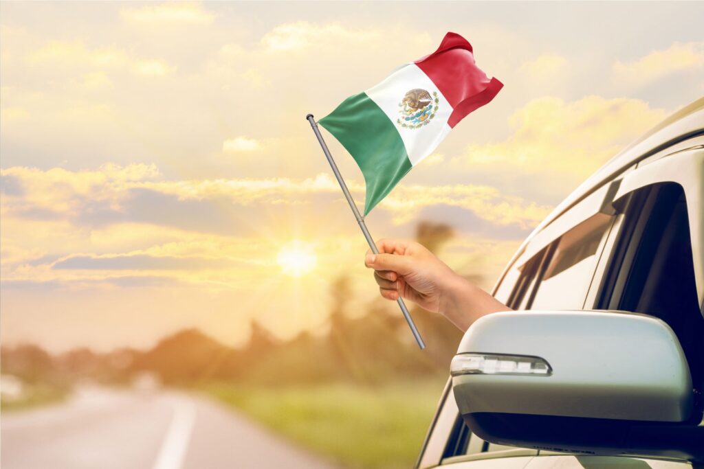 Grey car is in the bottom right corner, with a hand holding a small Mexican flag from the front passenger window. In the middle and on the left side, the sun is close to setting and is behind some clouds.