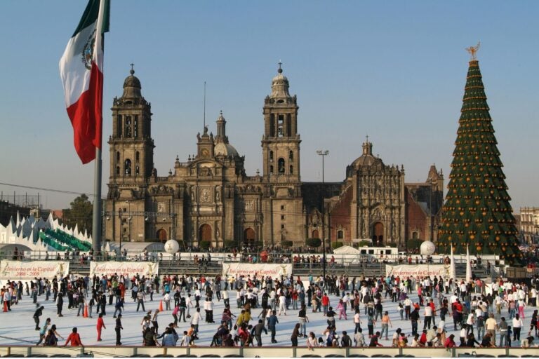 The plaza 'El Zocalo' in Mexico City. The Mexican flag on the left, a church in the middle and a Christmas tree on the right, with a temporary skating rink and people skating in front.