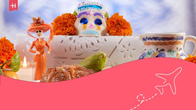 Table with traditional Mexican Catrina statue and sugar skull, among fruit and coffee cup. Holafly logo on the top left corner, and illustration of an aeroplane on the bottom right corner.