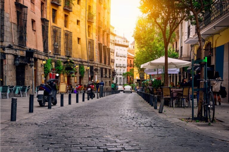 Cobble street in Madrid, Spain with people sitting at cafes and motorcycles and bicycles stationed along the sidewalk.