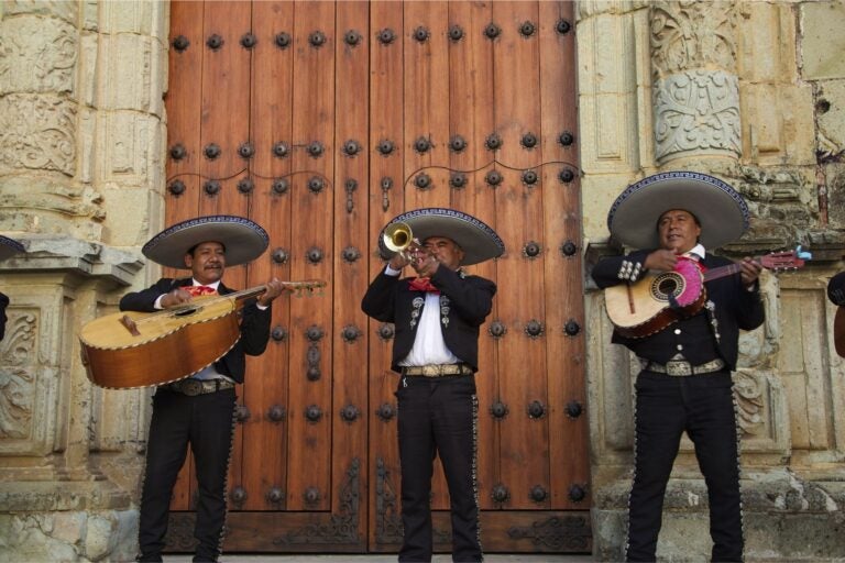 Three Mariachi band members playing their instruments in front of a large wooden door and colonial building.