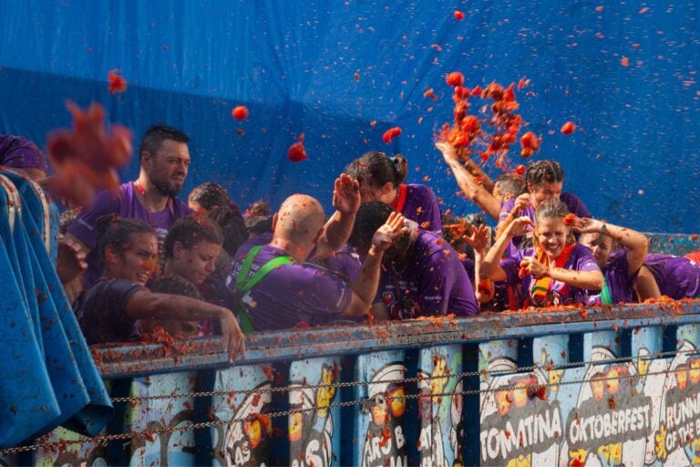 Participants of the Spanish festival La Tomatina laughing and throwing tomatoes in front of a large blue tarp.