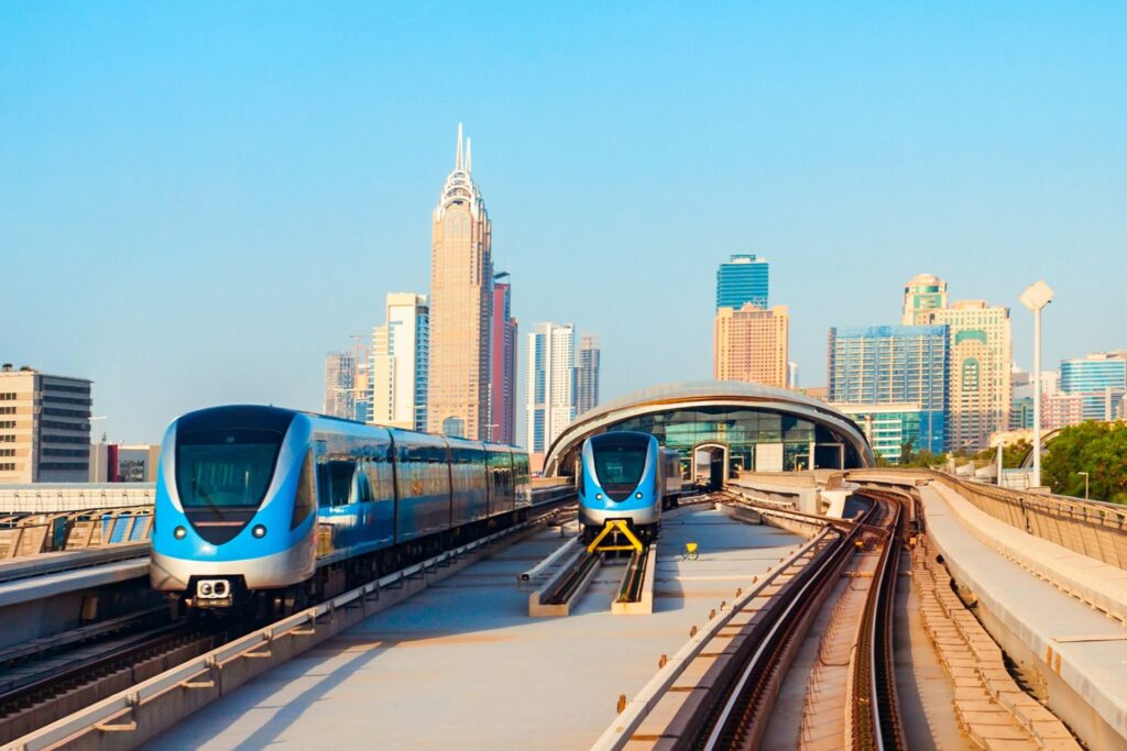 Front view of Dubai metro station during the day with two trains stationed next to one another. Dubai city skyline also seen in the distance.