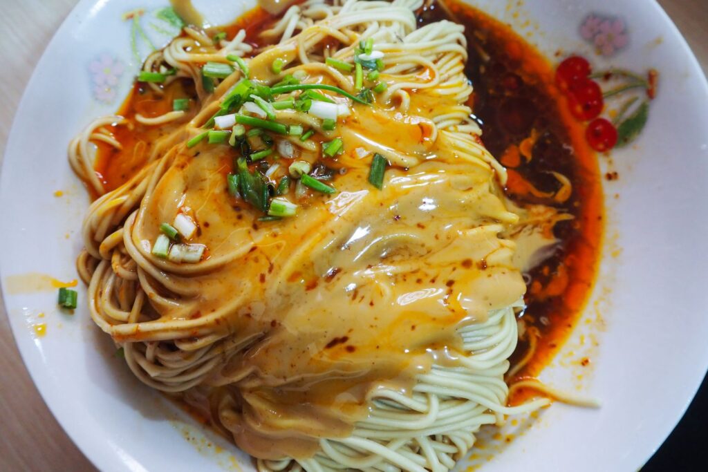 Noodles on a white plate with dark red scallion oil and sauce. Fresh green scallions on the top as a garnish.