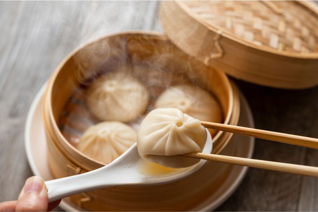 Close-up of the a person picking up one xiao long bao, or, soup dumpling from a bamboo steam bowl.