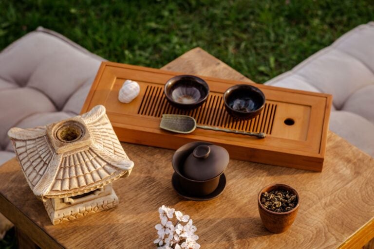 Wooden table on grass with beige cushions and traditional ceramic tea set on top of the table.