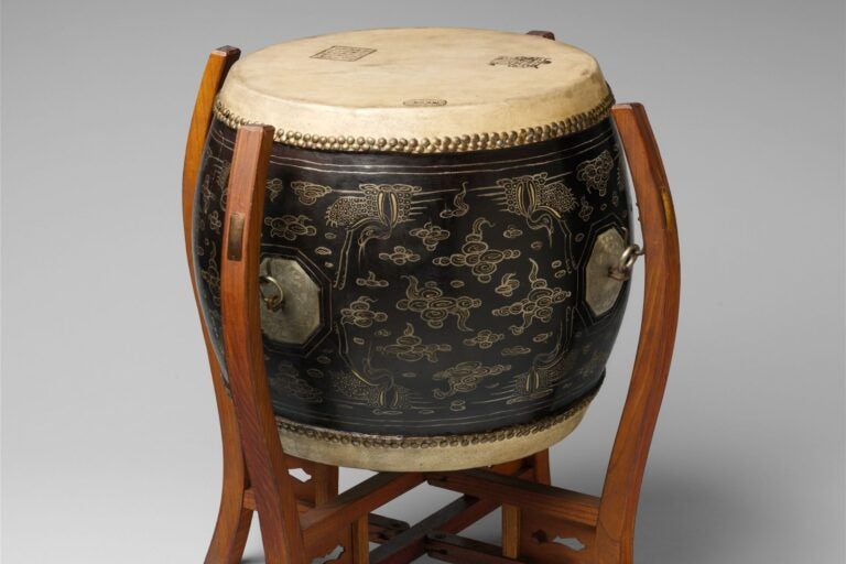 Black drum with gold painted designs, atop a wooden stand.