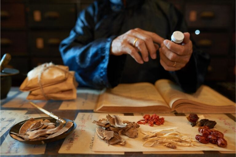 Chinese herbs atop papers with Chinese writing. Person holding a bottle of medicine in the background.

