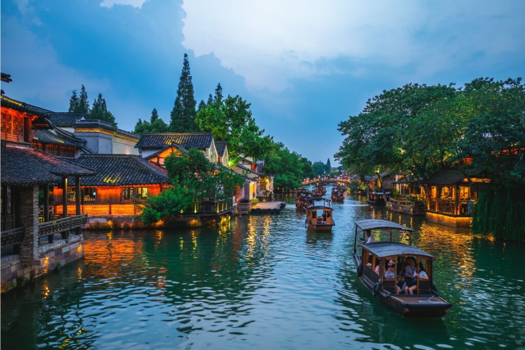The water town of Wuzhen in the evening with lit up buildings and boats sailing along the canal.