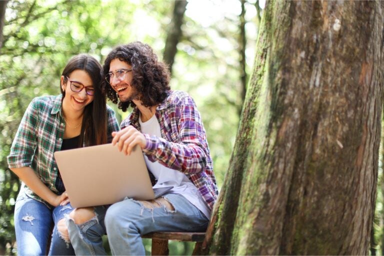 A young man and woman are sitting outside among trees, holding a laptop and laughing. 