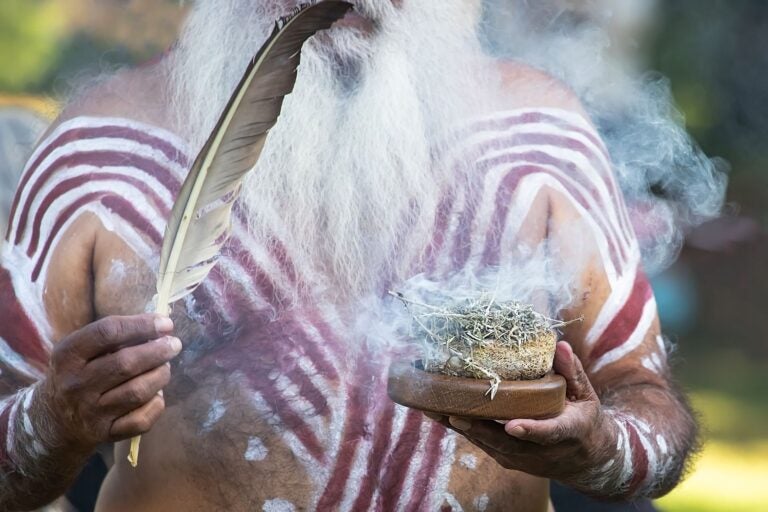 Close-up of an Indigenous Australian man with a long beard holding traditional smoke ceremony tools (feather and smoking materials).
