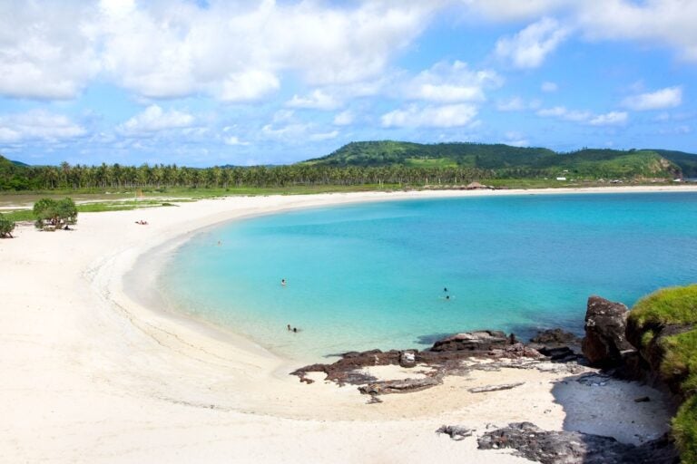 Side view of Tanjung Aan beach in Lombok, with people swimming in the ocean and palm trees and mountains in the background.