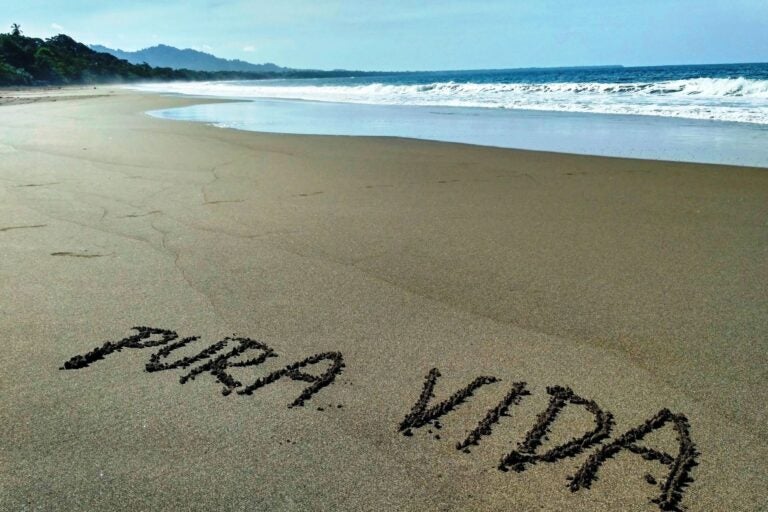 The words "Pura Vida" are written into the sand of a beach, with the shoreline and landscape in the background.