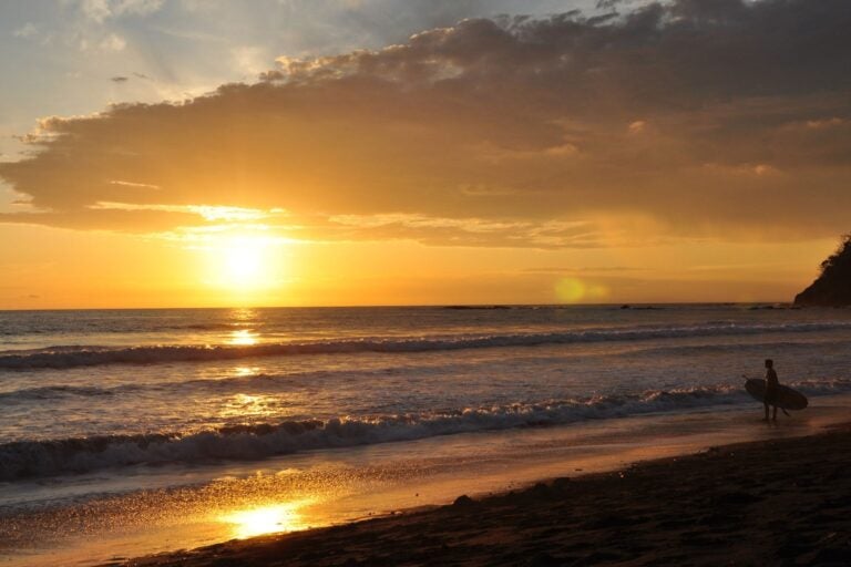 A beach at sunset with a man on the right side walking with a surf board.