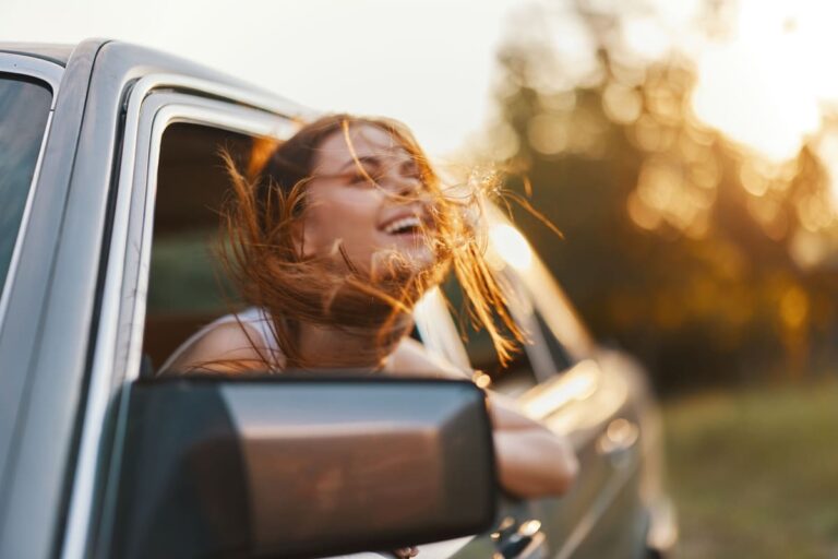 Joyful young woman enjoying a carefree moment with her hair blowing in the wind from a car window during a sunny day, embodying freedom and happiness.