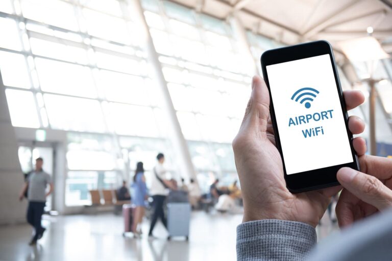 Traveler using a smartphone in an airport free Wi-Fi zone while passengers walk in the background.