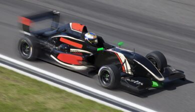 A black and red single-seat formula race car with a helmeted driver speeding along a paved circuit, motion blur emphasizing high speed on the track.