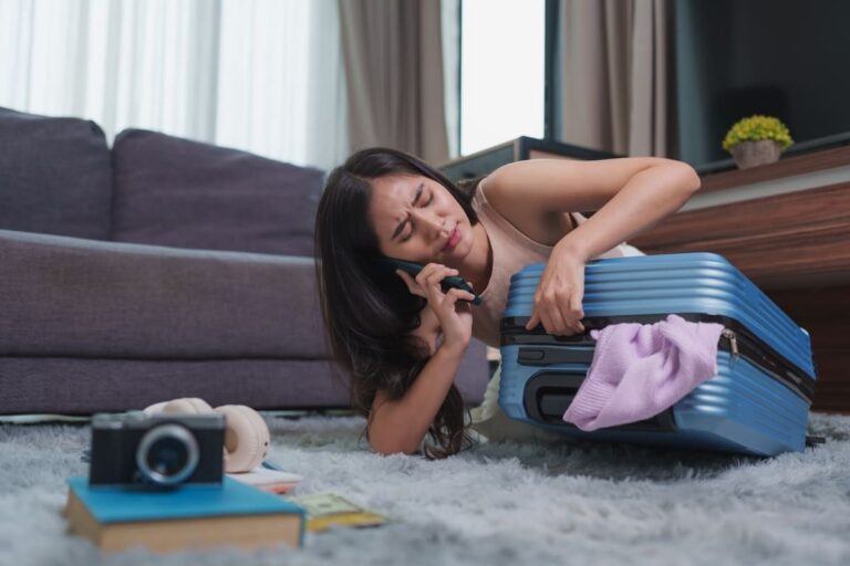 Young woman sitting on the floor trying to close an overpacked suitcase before a trip, looking stressed and overwhelmed.