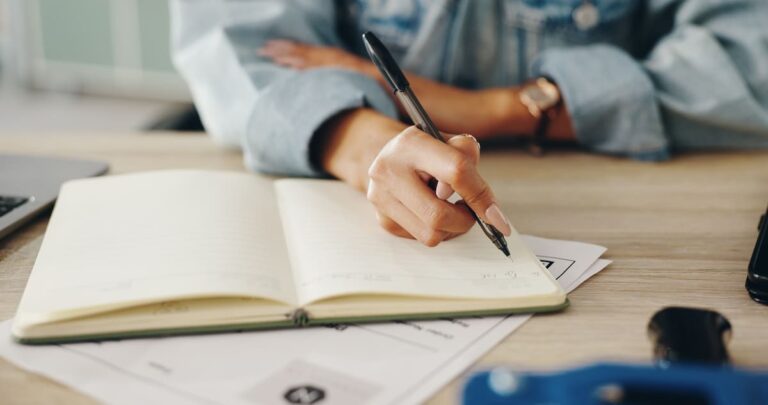 Woman writing in a notebook and reviewing a calendar while planning a travel itinerary at a desk.