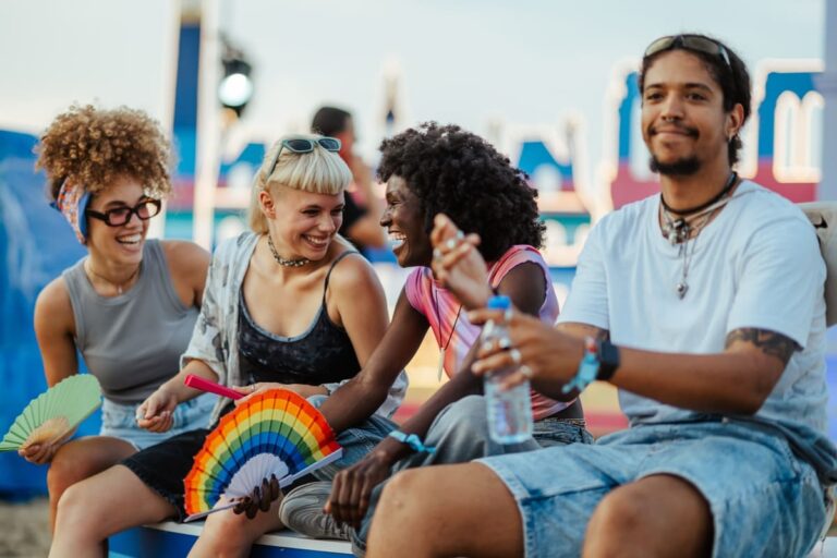 Four friends of different ethnicities sitting together at an outdoor music festival, holding colorful hand fans and a water bottle