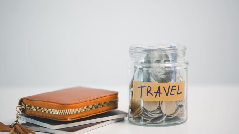 Glass jar labeled TRAVEL filled with coins and cash next to a wallet and notebooks