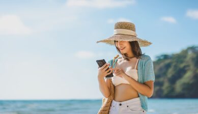 A young Asian woman relaxing on a sandy beach at Klong Chao Beach, Koh Kood, Thailand, smiling while using her mobile phone on a bright summer day.