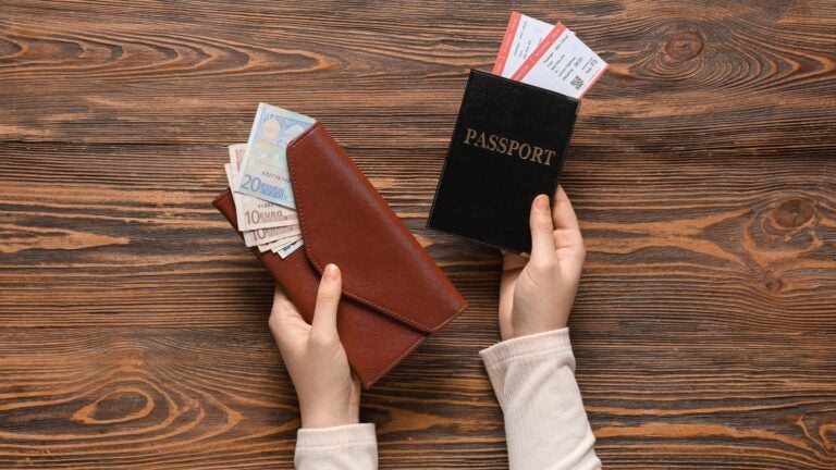 Close-up of a woman's hands holding an open wallet containing cash, a passport, and flight tickets on a wooden surface.