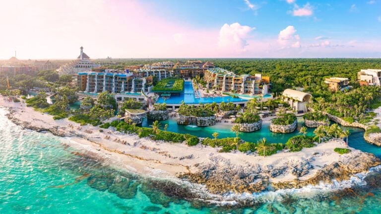 Aerial view of a large luxury resort complex bordering a white sand beach and turquoise Caribbean water in Cancun, Mexico, under clear sunny skies.