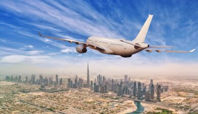 A commercial passenger aircraft flies over Dubai's downtown skyline, with high-rise buildings and urban development visible below.