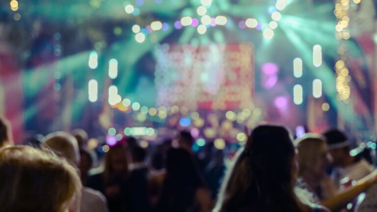 Silhouetted hands and figures of a crowd raised in the air against blurred, colorful lights at a nighttime music concert or festival.