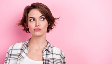 A young woman with a brown bob hairstyle looks pensively to one side with a skeptical expression, set against a bright pink background.