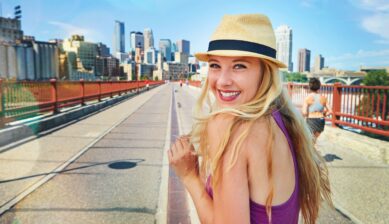 A smiling, excited young woman outdoors in a bustling urban area of Barcelona, with a blurred city street in the background, captured during a trip or vacation.