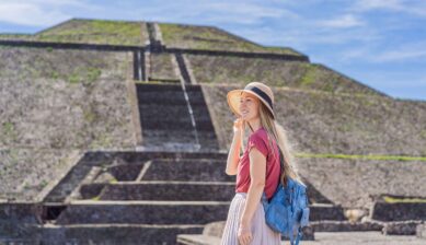 A female tourist stands among the ancient stone ruins at Teotihuacán, Mexico, with archaeological structures visible in the background under a bright sky.