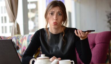 A stylish businesswoman sitting at a café table with a visibly frustrated expression, looking at her phone or laptop.