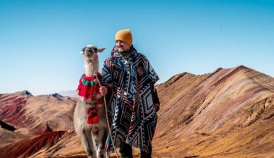 A man dressed in traditional Peruvian clothing stands alongside a llama on the vivid red and pink striped slopes of Rainbow Mountain under a bright blue sky.