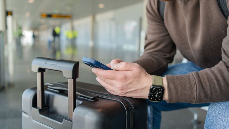 A smiling man sits in an airport terminal with a travel bag beside him, looking at his mobile phone while waiting for his flight.