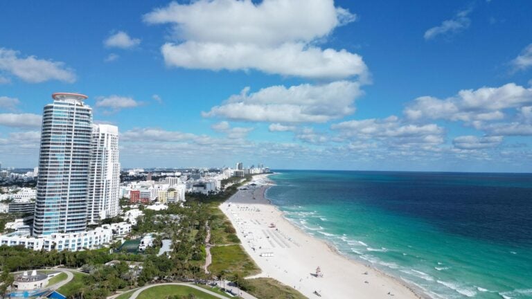 An aerial panoramic view of Miami's South Beach waterfront, showing the marina, high-rise buildings along the coastline, and the blue waters of the Atlantic Ocean stretching into the distance.