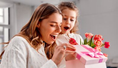 Happy mother and daughter opening a pink gift box together with a bouquet of tulips at home on Mother's Day