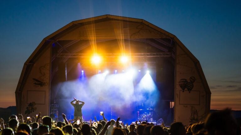 An outdoor festival stage lit with spotlights and smoke as a crowd watches after dark