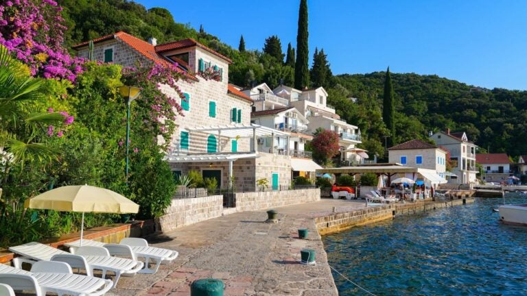 A calm waterfront promenade lines a small stone fishing village on the Luštica Peninsula, with traditional Montenegrin architecture reflected in the still waters of the Adriatic Sea.