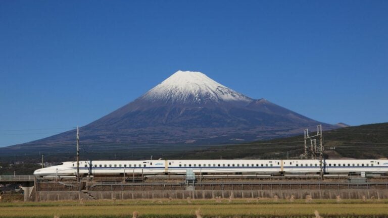 A white shinkansen bullet train passing green fields with Mount Fuji behind