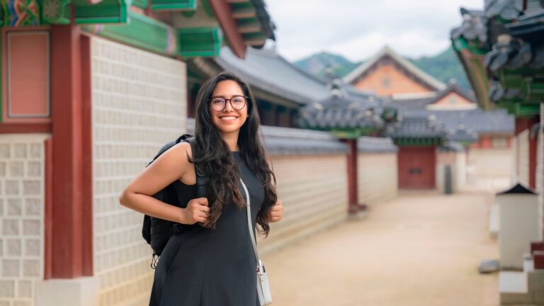  A young Latina woman smiling in the sunlit courtyard of Gyeongbokgung Palace in Seoul, South Korea, with traditional temple architecture visible behind her.