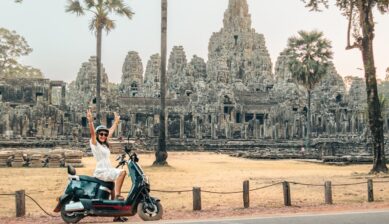 A smiling young Caucasian traveler sits on a rented black electric scooter in front of the ancient stone towers of Angkor Wat, with the temple's iconic silhouette rising behind him against a bright sky.