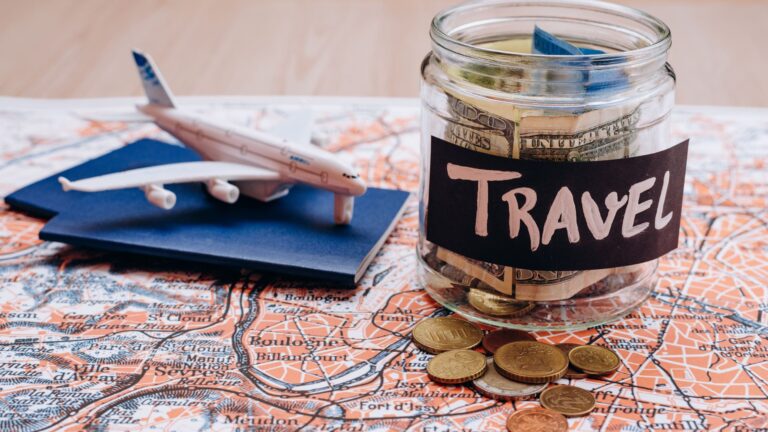 A glass jar filled with coins and paper currency sits in front of a world map, with a small label reading "travel budget" — a visual representation of saving money for an international trip.