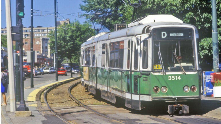 A classic red and cream trolley car travels along a street in Boston, Massachusetts, with period architecture and urban streetscape visible in the background.