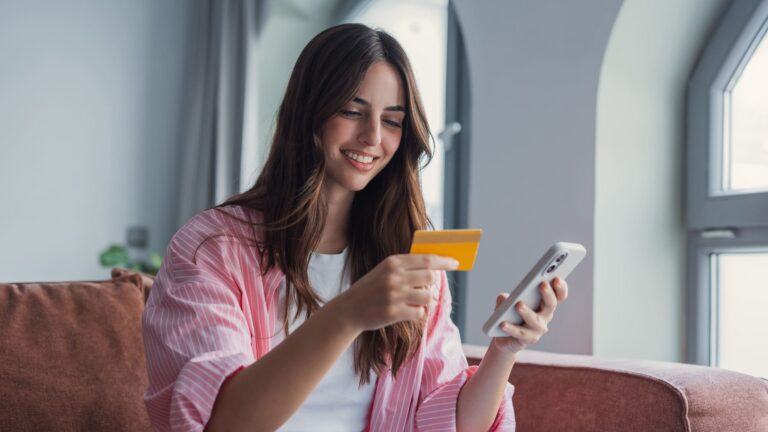 A smiling Latina woman holding a credit card and smartphone while making an online payment from her home.