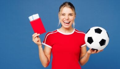 A woman dressed in red holds a passport, match ticket, and soccer ball against a plain blue background, representing an international football fan preparing to travel abroad for a tournament.