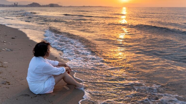 A woman sits cross-legged on a sandy beach at sunrise, eyes closed in meditation, with calm water and soft golden light in the background.