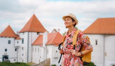 A woman walks through the historic old town of Varaždin in Croatia on a sunny summer day, with cobblestone streets and well-preserved baroque buildings lining the pedestrian area behind her.